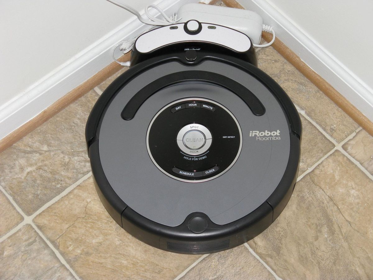 Grau-schwarzer iRobot Roomba mit Bedientasten auf der Oberseite steht an seiner Ladestation in der Ecke eines Raums mit Fliesenboden
