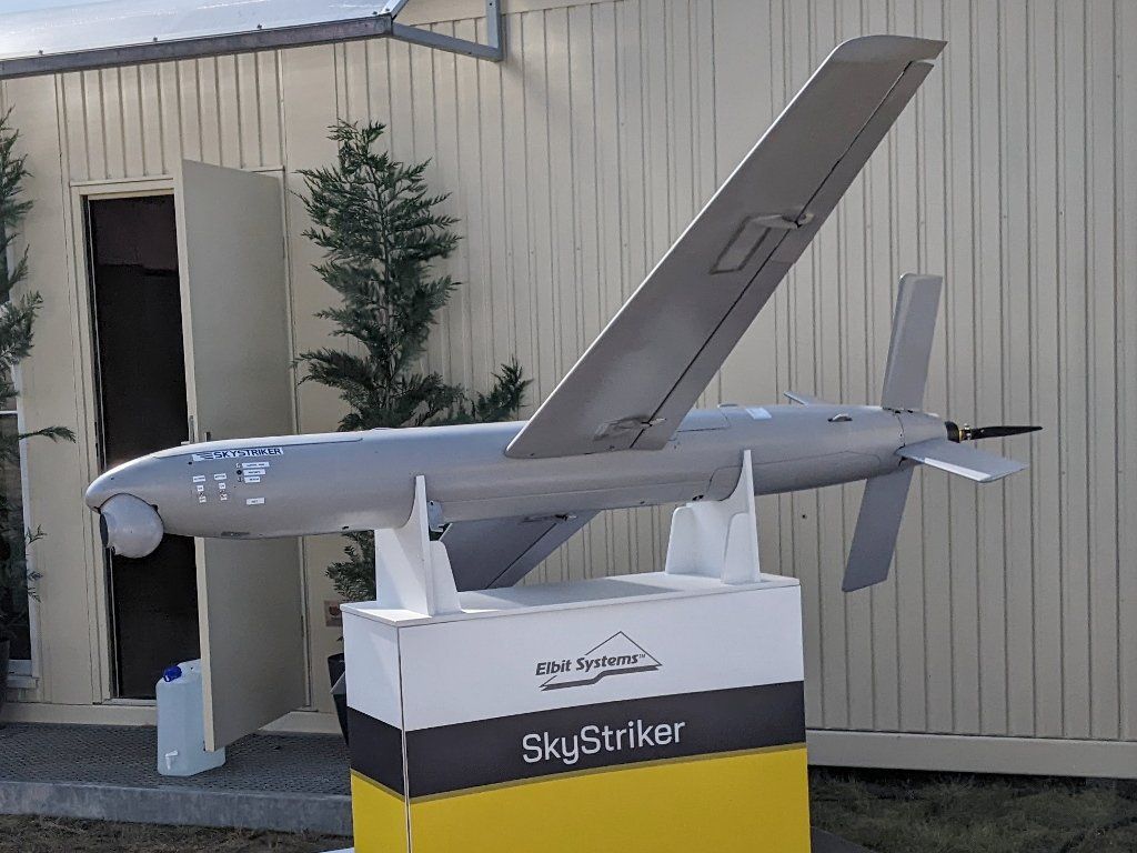 Hellgrauer, schlanker Flugkörper mit schräg gestellter Tragfläche, Kreuzleitwerk, Heckpropeller und Kamerakugel am Bug auf zwei Halterungen über einem Sockel mit der Aufschrift Elbit Systems SkyStriker, im Hintergrund eine Containerwand und eine kleine Konifere
