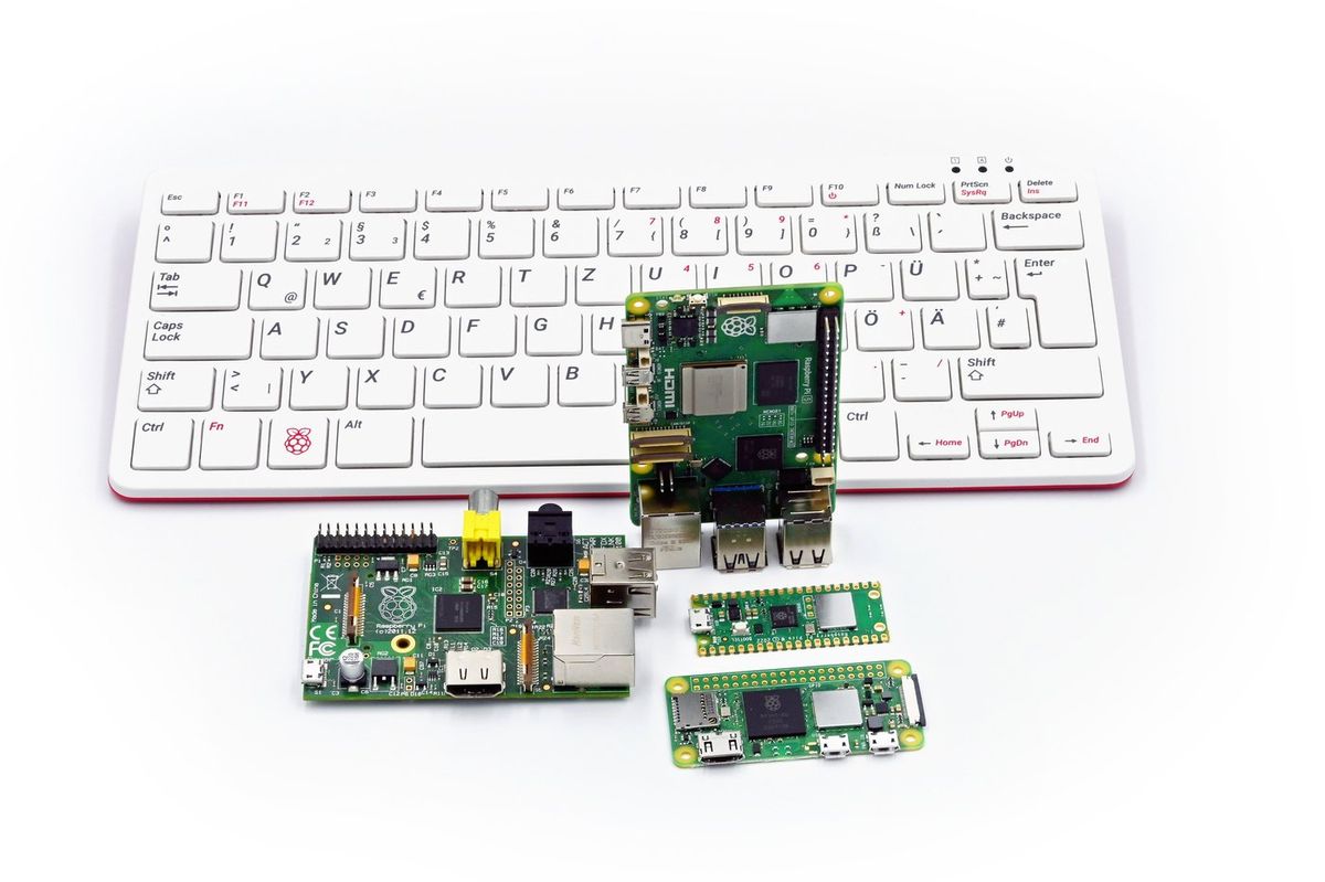 Weiße Raspberry-Pi-Tastatur mit deutschem Layout, davor eine große grüne Platine der ersten Generation, ein aufrecht stehender Raspberry Pi 5 sowie zwei schmale Platinen: ein Pico und ein Zero 2