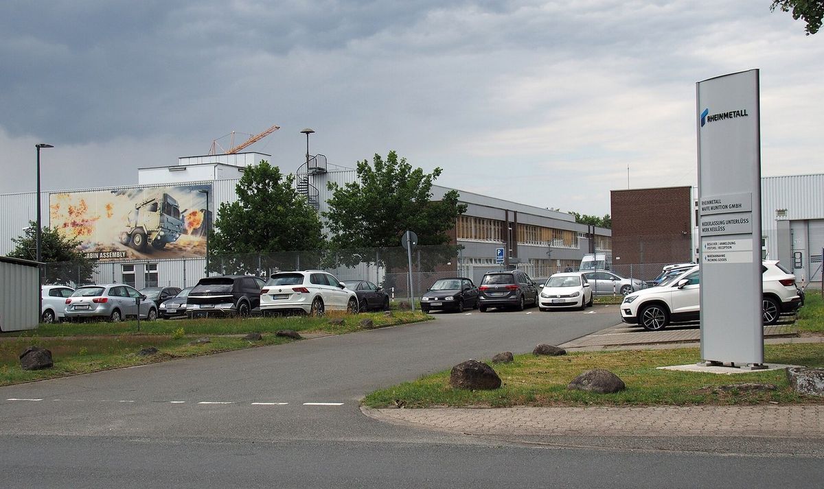 Hallen eines Industriewerks mit Parkplatz unter grauem Himmel, rechts eine Stele mit der Aufschrift Rheinmetall Waffe Munition GmbH, Niederlassung Unterlüss, Werk Neulüss, links ein großes Werbebild eines Militärlastwagens
