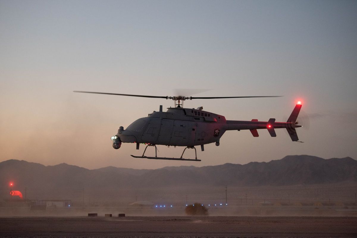 Grauer unbemannter Hubschrauber mit Kufen, Kamerakugel unter dem Bug und roten Positionslichtern schwebt in der Abenddämmerung knapp über staubigem Wüstenboden, dahinter Berge und blaue Lichter einer Landefläche