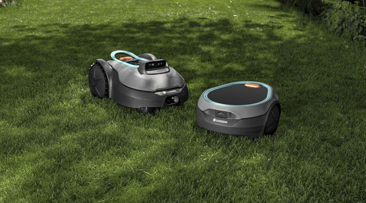 Zwei graue Mähroboter auf einem sonnigen, teils beschatteten Rasen: links ein größeres Modell mit türkisfarbenem Tragebügel und Kameraaufsatz, rechts ein flaches, rundliches Modell mit dunkler Solarfläche auf der Oberseite