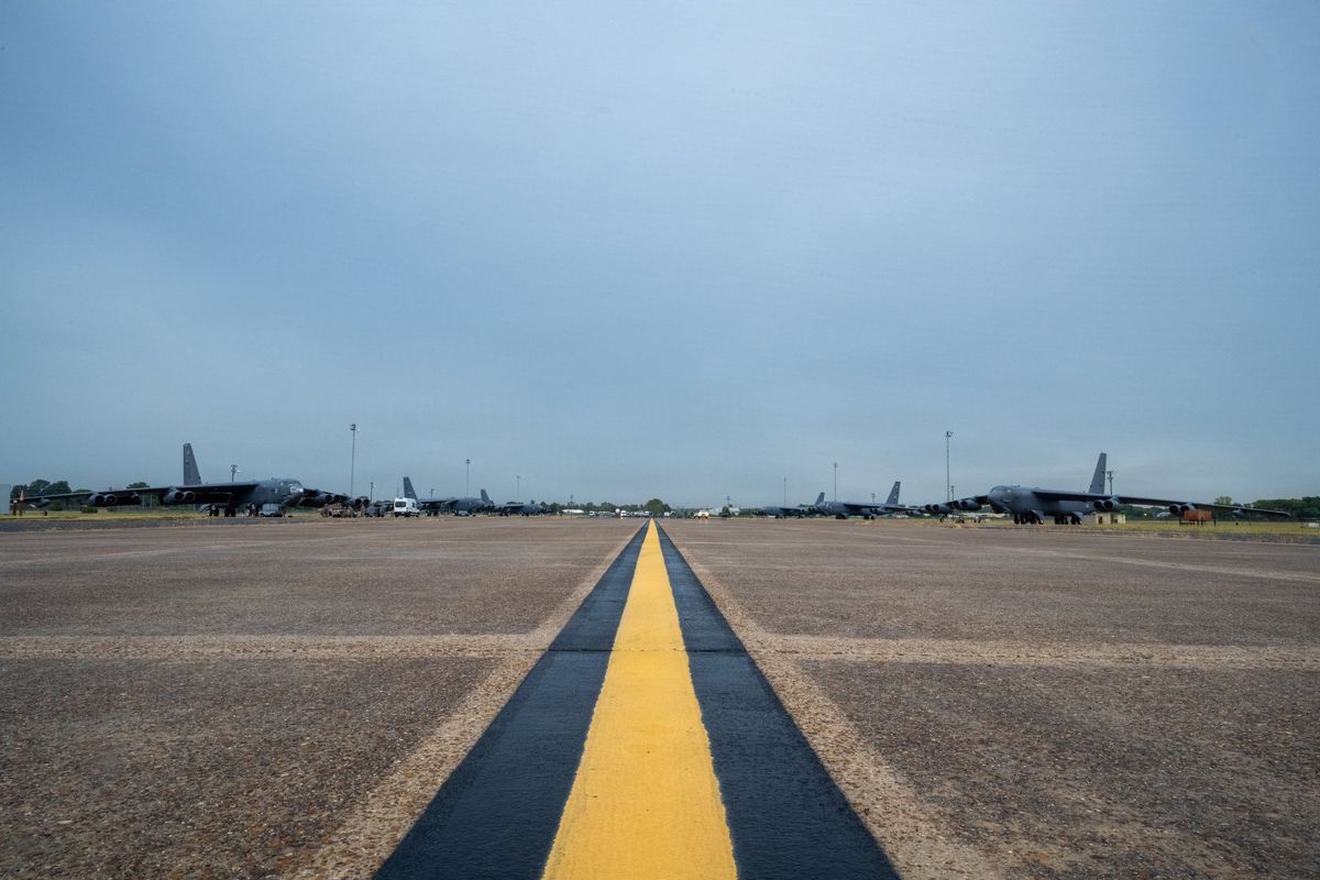 Blick entlang einer gelben Mittellinie über ein weites Vorfeld, am Horizont stehen links und rechts sechs B-52H-Bomber, dazwischen Fahrzeuge und Flutlichtmasten, darüber ein grauer Himmel