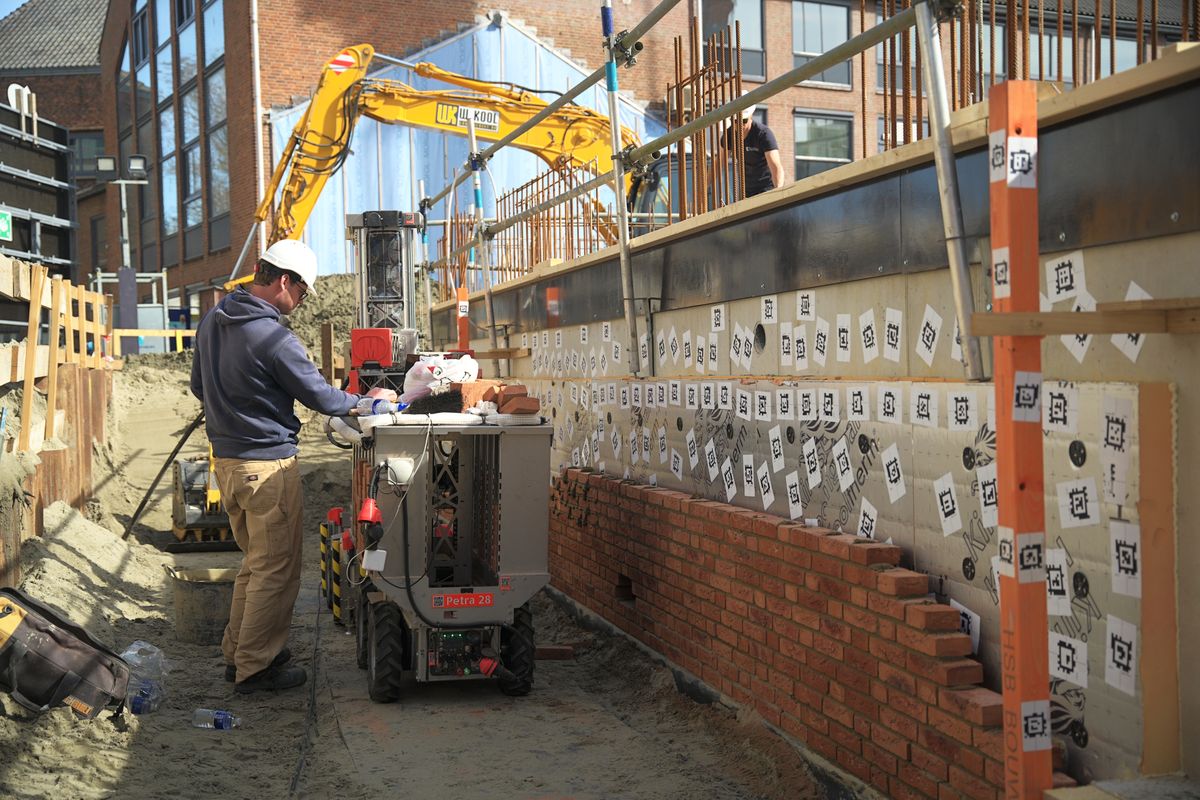 Monumental-Mitarbeiter bedient einen Roboter an einer entstehenden Mauer, im Hintergrund ein Bagger