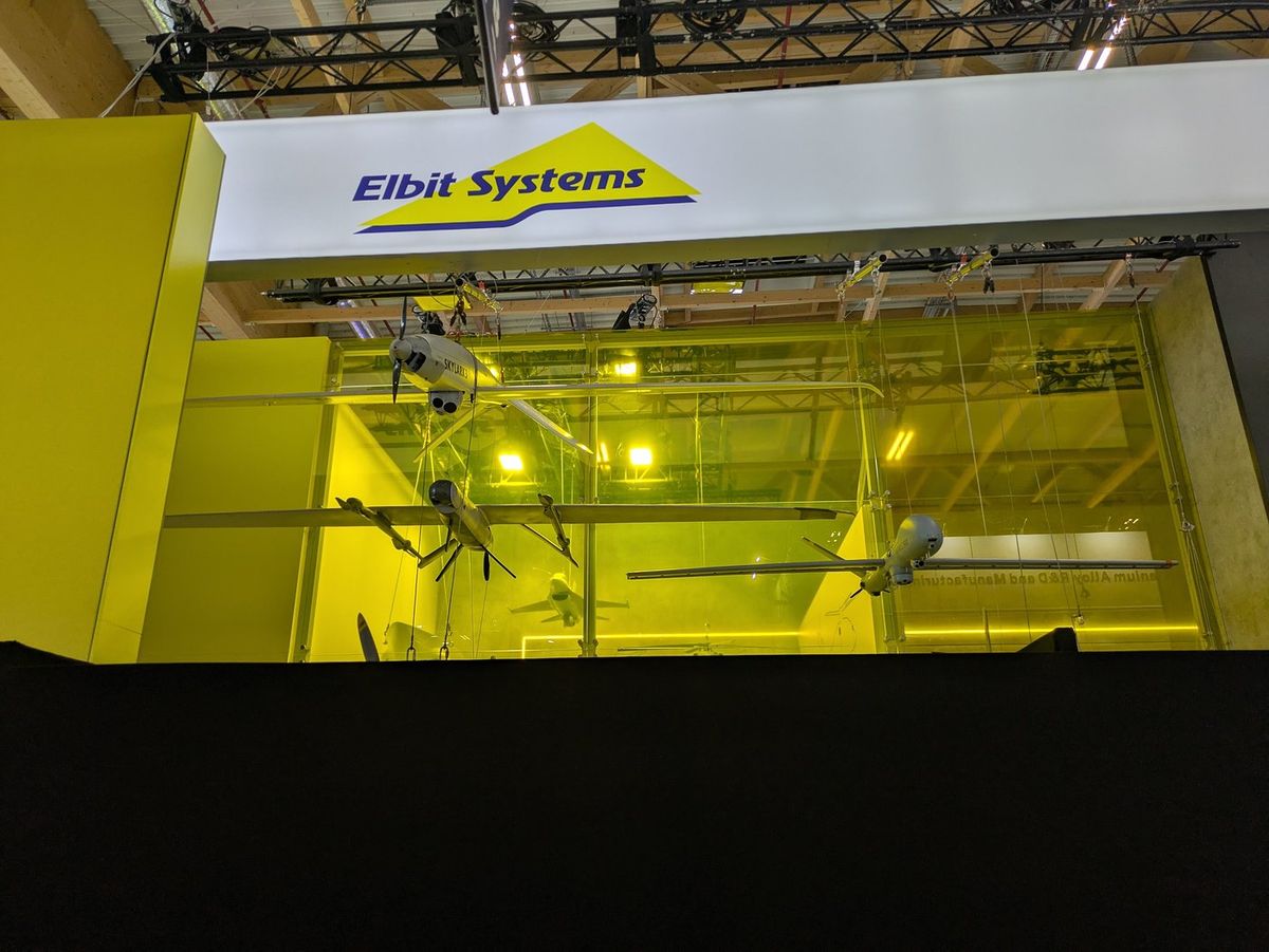 Messestand mit gelben Wänden und einem weißen Banner mit dem Schriftzug Elbit Systems, darunter hängen Drohnenmodelle, darunter eine Skylark 3, vor einer gelben Glaswand; die untere Hälfte des Stands ist mit einer schwarzen Wand verdeckt
