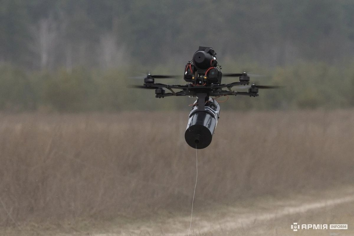 Fliegende Quadrocopter-Drohne mit einem großen zylindrischen Sprengkörper unter dem Rumpf, von dem ein dünner Faden senkrecht nach unten hängt