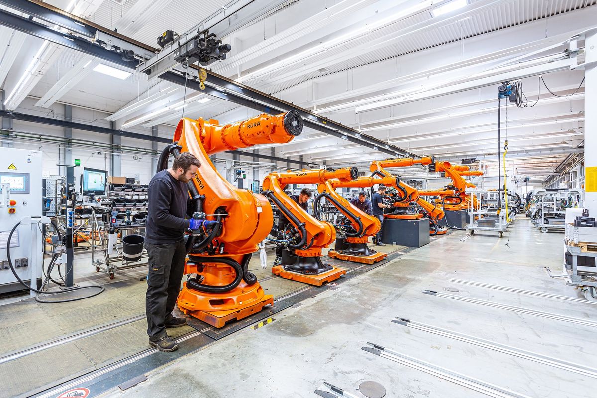 Montagehalle mit einer Reihe großer orangefarbener KUKA-Industrieroboter auf Montageplätzen, im Vordergrund arbeitet ein Mitarbeiter am Sockel eines Roboters, darüber Hallenkräne
