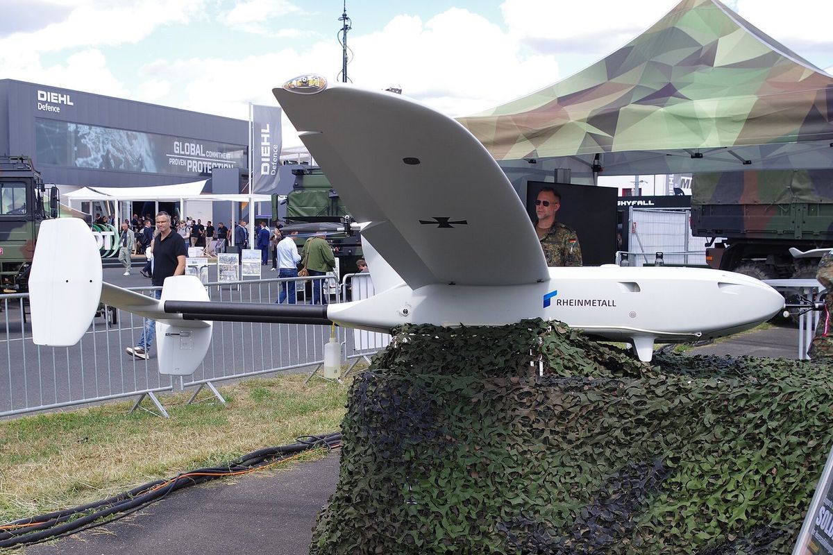Weiße Aufklärungsdrohne mit langem, schlankem Flügel, doppeltem Leitwerksträger, Bundeswehr-Kreuz und Rheinmetall-Schriftzug am Rumpf steht auf einem mit Tarnnetz bedeckten Sockel, im Hintergrund Messebesucher, ein Soldat und Stände von Diehl Defence