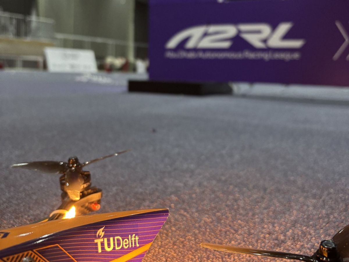 Nahaufnahme einer Renndrohne mit orange-violetter Verkleidung und TU-Delft-Logo auf grauem Teppichboden, die vordere Haube im Bildvordergrund, links ein Propeller mit rot und orange leuchtenden LEDs, dahinter unscharf ein violettes Banner mit der Aufschrift A2RL Abu Dhabi Autonomous Racing League