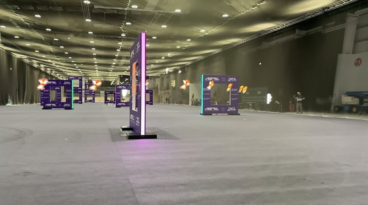 Blick durch eine lange Messehalle mit grauem Teppichboden und Lichterreihen an der Decke; violette Renntore mit A2RL- und DCL-Logos stehen verteilt im Raum, das vorderste mit violett leuchtenden Kanten, und an mehreren Toren hängen orange leuchtende Lichtspuren einer Drohne in der Luft; rechts am Rand zwei Personen und eine Hebebühne