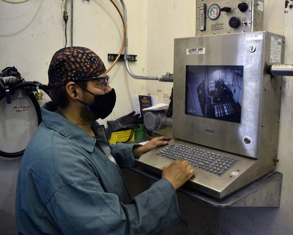 Ein Beschäftigter sitzt an einem Bedienpult mit Tastatur und Monitor und beobachtet auf dem Bildschirm einen Roboterarm in einem Nebenraum
