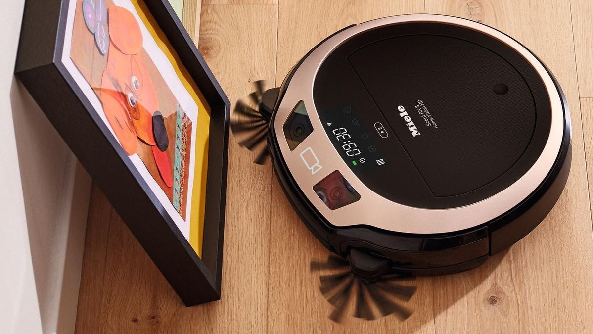 Der roségoldene Miele Scout RX3 Home Vision HD von schräg oben mit Display, zwei Frontkameras und rotierenden Seitenbürsten an einem Bilderrahmen.