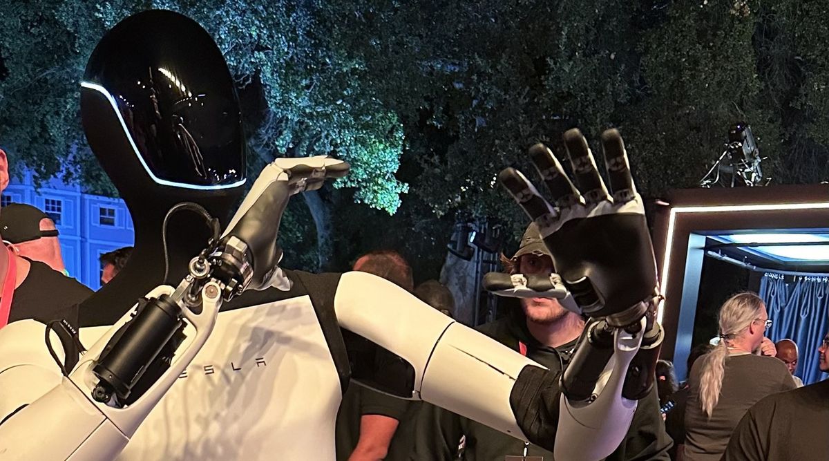 Weißer humanoider Roboter Optimus mit schwarz glänzendem Kopf und TESLA-Schriftzug auf der Brust hebt bei einer abendlichen Veranstaltung unter Bäumen eine Hand mit gespreizten schwarz-weißen Fingern, dahinter Besucher und ein beleuchteter Pavillon