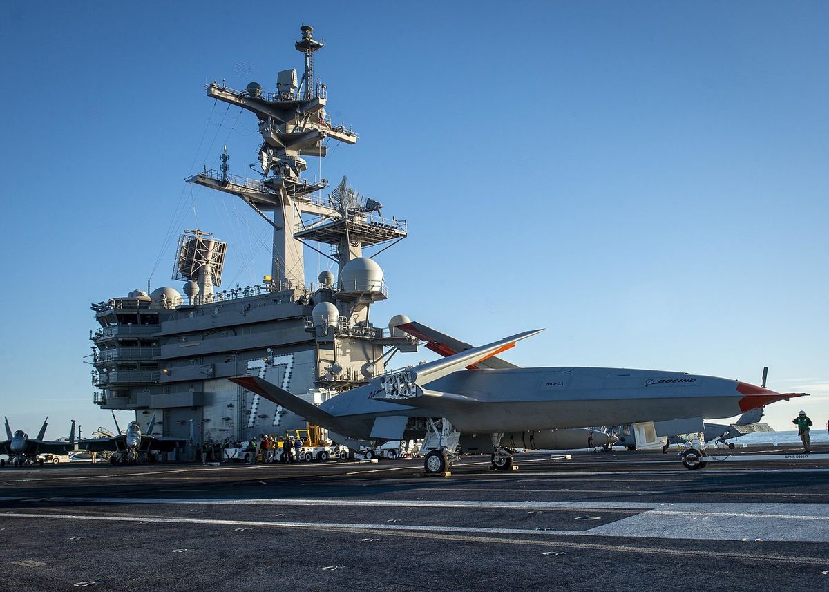Graue Drohne ohne Cockpit mit roter Nasenspitze, nach oben geklappten Flügelspitzen und der Aufschrift MQ-25 und Boeing steht auf dem Flugdeck eines Flugzeugträgers, dahinter die Kommandoinsel mit der Nummer 77 und Antennenmasten, links geparkte Kampfjets, rechts ein Deckarbeiter mit Helm
