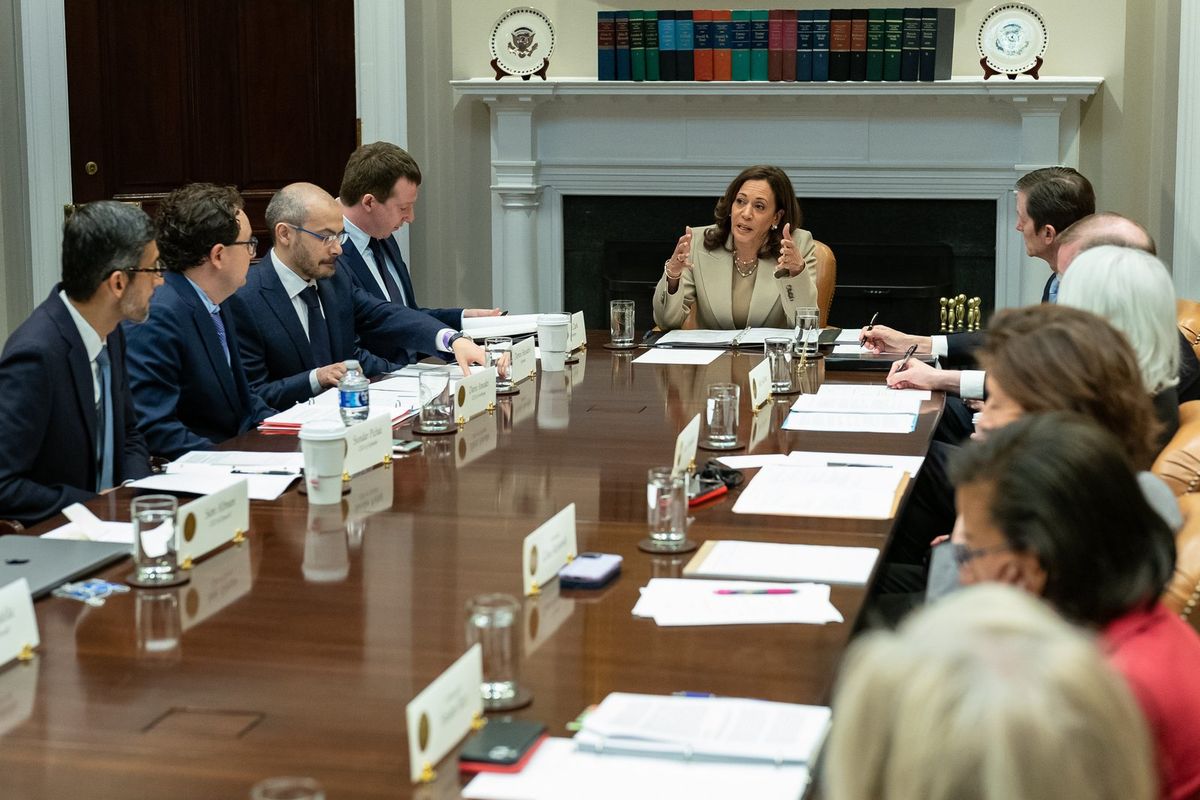 Kamala Harris spricht gestikulierend am Kopfende eines langen Holztischs vor einem weißen Kamin mit Buchreihe, links vier Männer in dunklen Anzügen mit Namensschildern und Unterlagen, rechts Teilnehmer von hinten