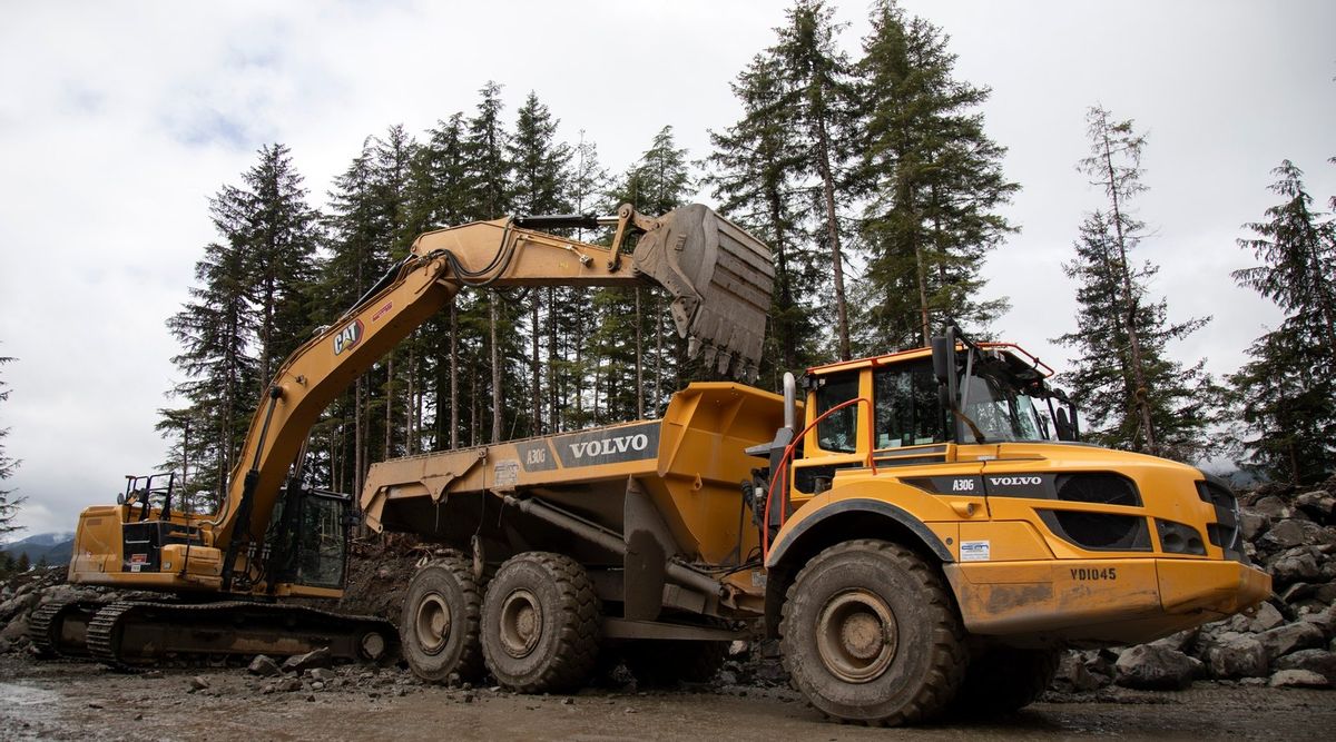 Ein gelber Kettenbagger mit CAT-Logo hebt seine gefüllte Schaufel über die Mulde eines gelben Volvo-Knickgelenk-Dumpers A30G, davor schlammiger Boden und Felsbrocken, dahinter hohe Nadelbäume unter bewölktem Himmel