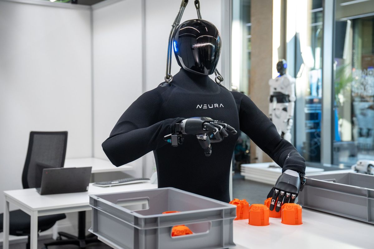 Humanoider Roboter in schwarzem Textilanzug mit NEURA-Schriftzug und glänzend schwarzem Helmkopf, an Gurten aufgehängt, greift mit einer schwarzen Roboterhand nach orangefarbenen Kunststoffteilen auf einem weißen Tisch; davor eine graue Kunststoffkiste, links ein Schreibtisch mit Laptop und Bürostuhl, im Hintergrund ein zweiter weißer Humanoid hinter einer Glaswand