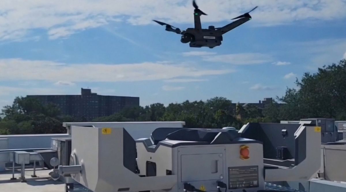 Eine dunkelgraue Quadrokopter-Drohne schwebt über einer hellgrauen Dockingstation mit seitlich aufgeklappter Abdeckung auf einem Flachdach, im Hintergrund Bäume und ein Hochhaus unter blauem Himmel