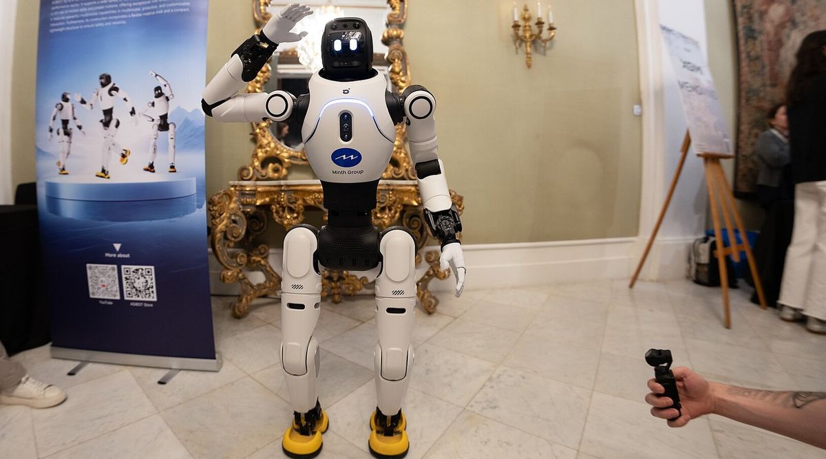 Weißer humanoider Roboter AgiBot X2 mit gelben Füßen hebt grüßend die rechte Hand, auf der Brust ein blaues Logo mit der Aufschrift Minth Group, links ein blauer Roll-up-Banner mit drei Abbildungen des Roboters, dahinter ein vergoldeter Barockspiegel auf einer Marmorkonsole, rechts eine Hand mit einer kleinen Kamera