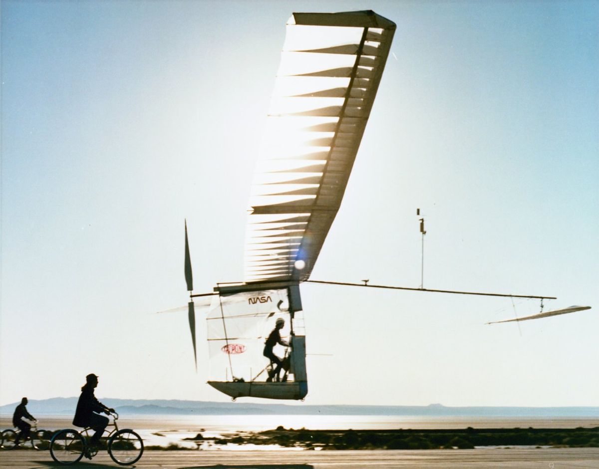 Ein Muskelkraftflugzeug mit langem, durchscheinendem Flügel und Propeller am Heck schwebt im Gegenlicht knapp über einer Piste, in der offenen Kabine tritt ein Pilot in die Pedale, vorne fahren zwei Radfahrer mit