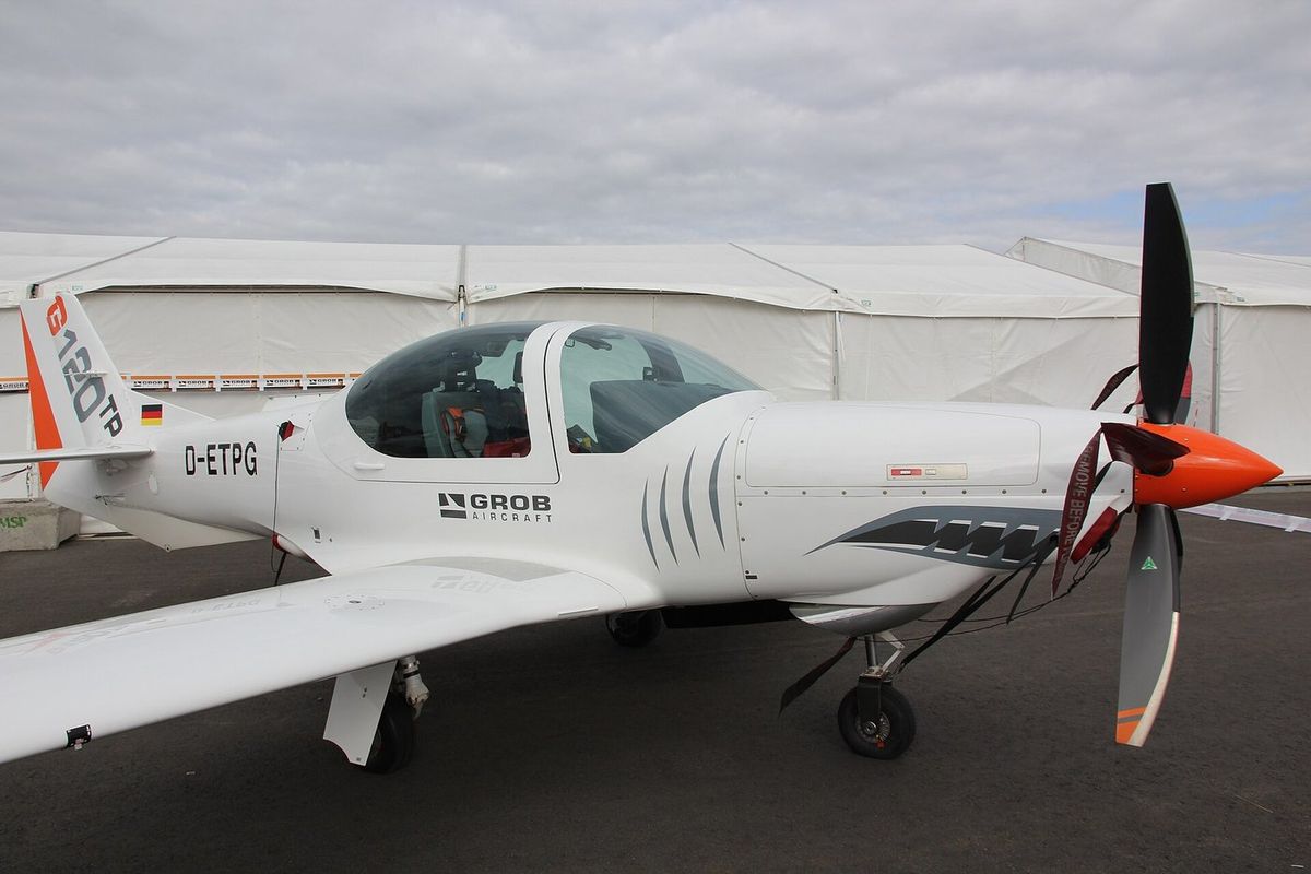 Weißes einmotoriges Propellerflugzeug mit zweisitziger Glaskanzel, orangefarbener Propellerhaube, dunklen Propellerblättern, Kennzeichen D-ETPG und Grob-Aircraft-Schriftzug am Rumpf steht auf Asphalt vor weißen Messezelten