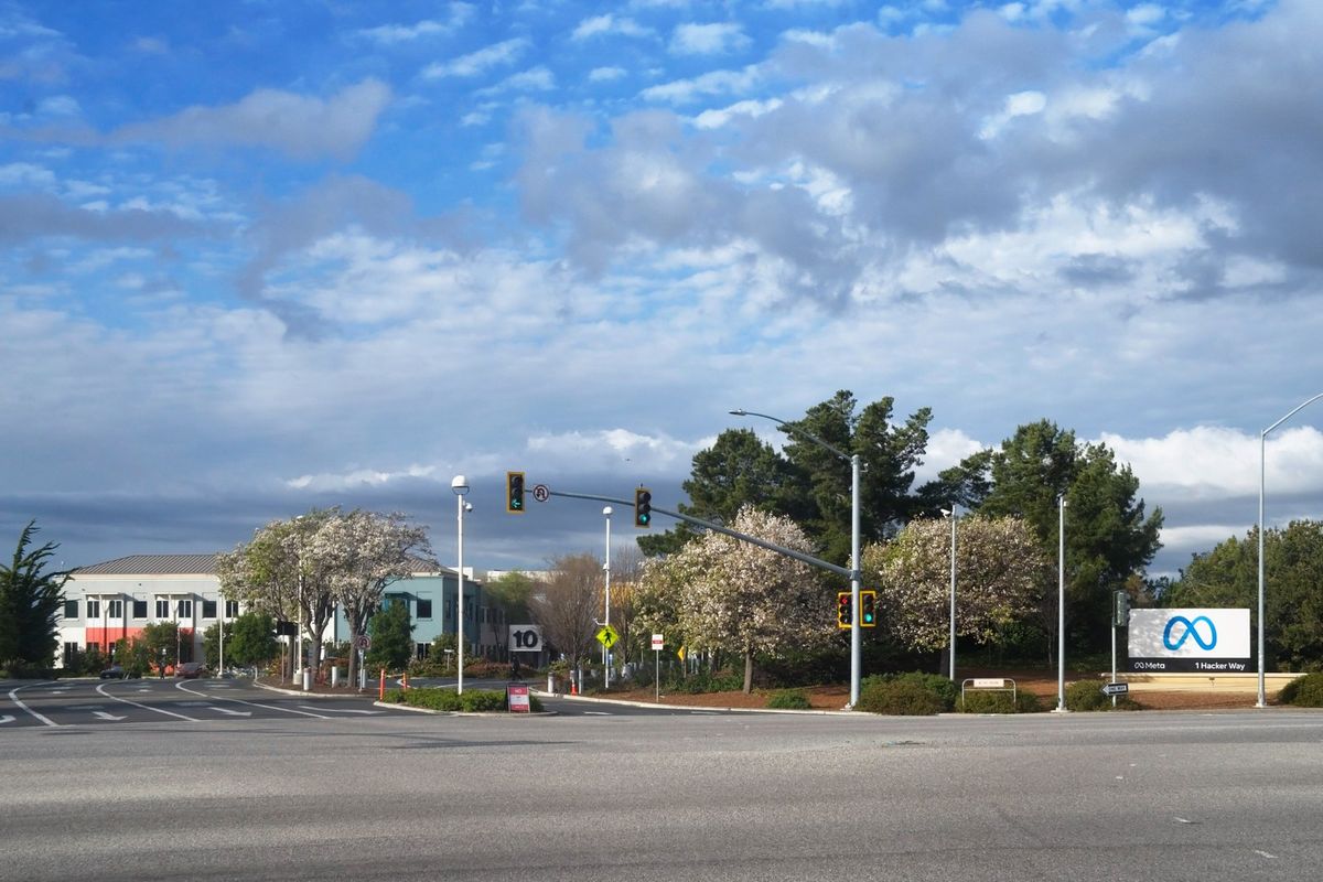 Weite Straßenkreuzung mit Ampeln unter bewölktem Himmel, rechts ein weißes Schild mit blauem Unendlichkeitszeichen und der Aufschrift Meta und 1 Hacker Way, dahinter blühende Bäume und ein helles Bürogebäude mit der Nummer 10
