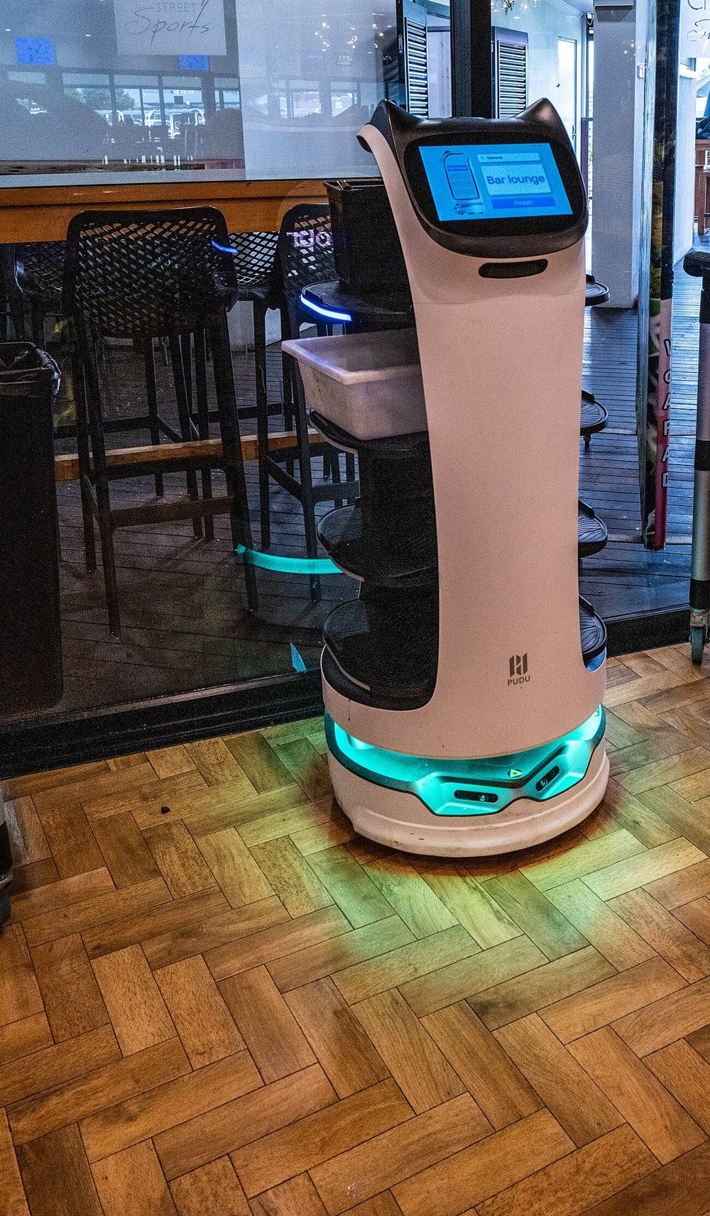 Weißer Servierroboter Pudu BellaBot mit Katzenohren und Touch-Display am Kopf, Pudu-Logo am Korpus und türkis leuchtendem Fahrgestell auf Fischgrätparkett.