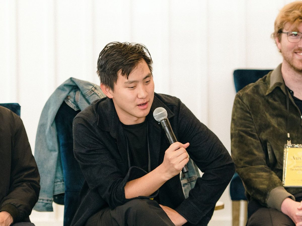 Alexandr Wang in schwarzer Jacke und schwarzem T-Shirt sitzt vorgebeugt auf einem Stuhl und spricht in ein Handmikrofon, über der Lehne hinter ihm hängt eine Jeansjacke, rechts ein Mann mit rotblonden Haaren, Brille und Namensschild, hinten eine weiße Holzwand