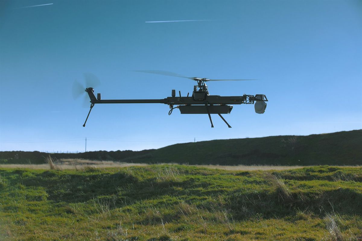 Schlanke schwarze Hubschrauberdrohne mit langem Ausleger und Heckrotor schwebt knapp über einer Wiese vor blauem Himmel