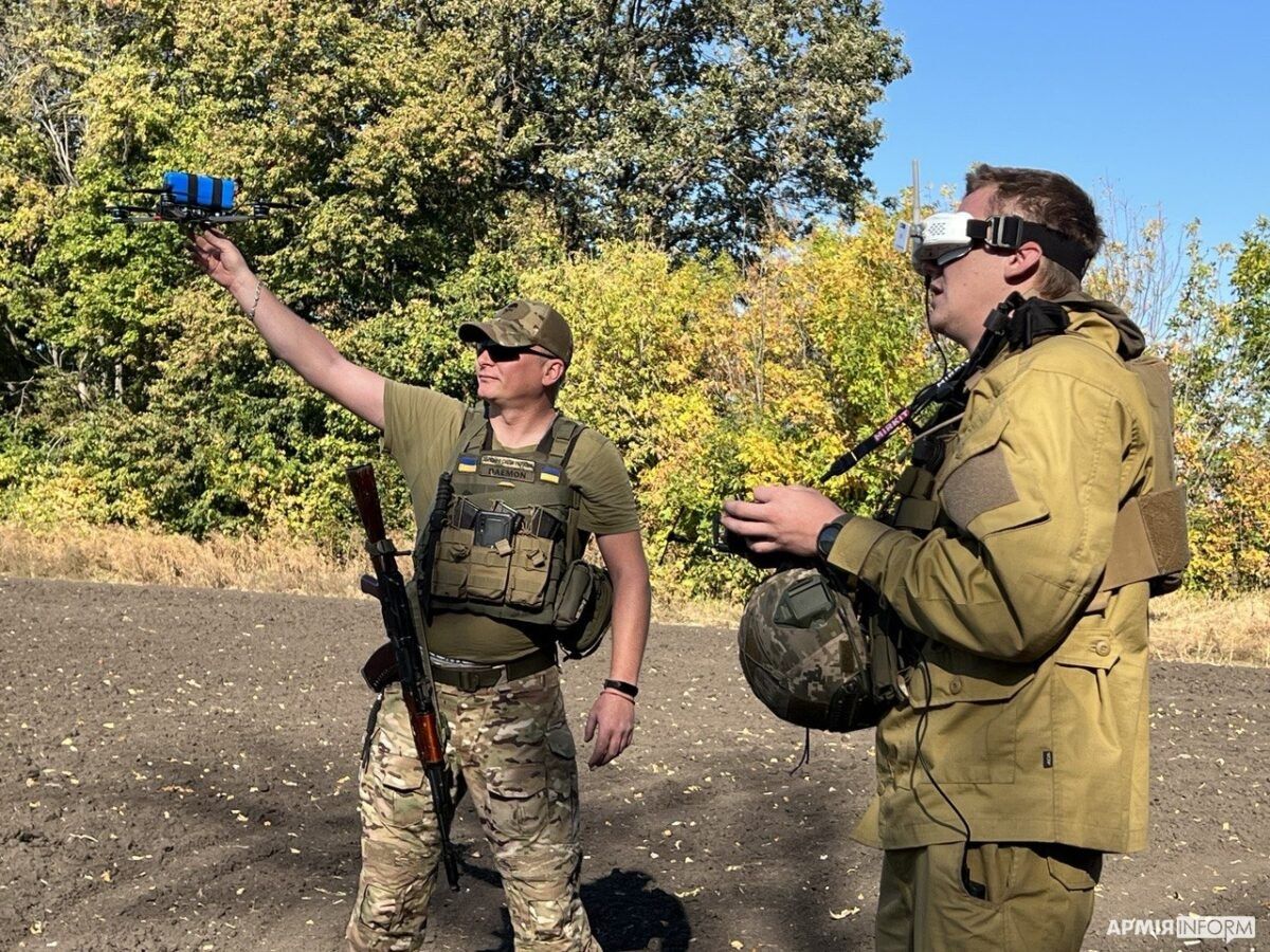Ein Soldat hält eine kleine Quadrocopter-Drohne mit einem blauen Sprengsatz in die Höhe, daneben steuert ein zweiter Soldat mit Videobrille und Fernsteuerung
