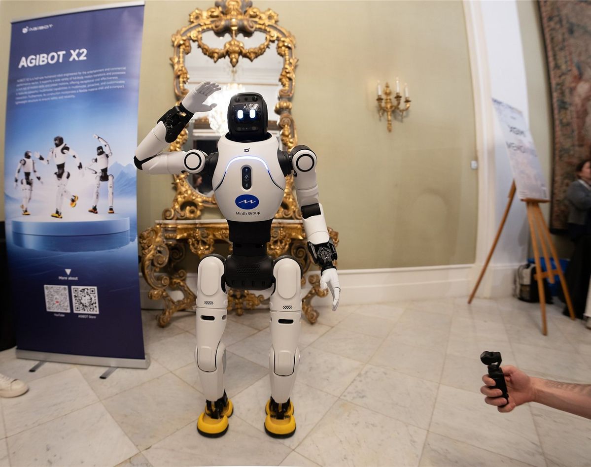 Weißer humanoider Roboter AgiBot X2 hebt grüßend die rechte Hand, im Hintergrund ein Roll-up mit der Aufschrift AGIBOT X2 und ein vergoldeter Spiegel.