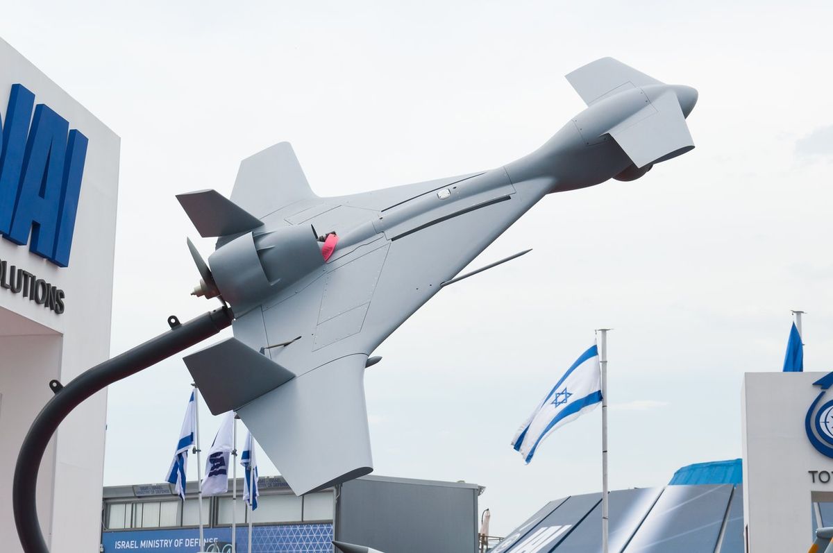 Hellgraues Fluggerät mit Deltaflügeln, kleinen Entenflügeln am Bug und Heckpropeller ist schräg nach oben auf einem gebogenen Mast montiert, links ein Messestand mit blauem IAI-Schriftzug, rechts israelische Flaggen