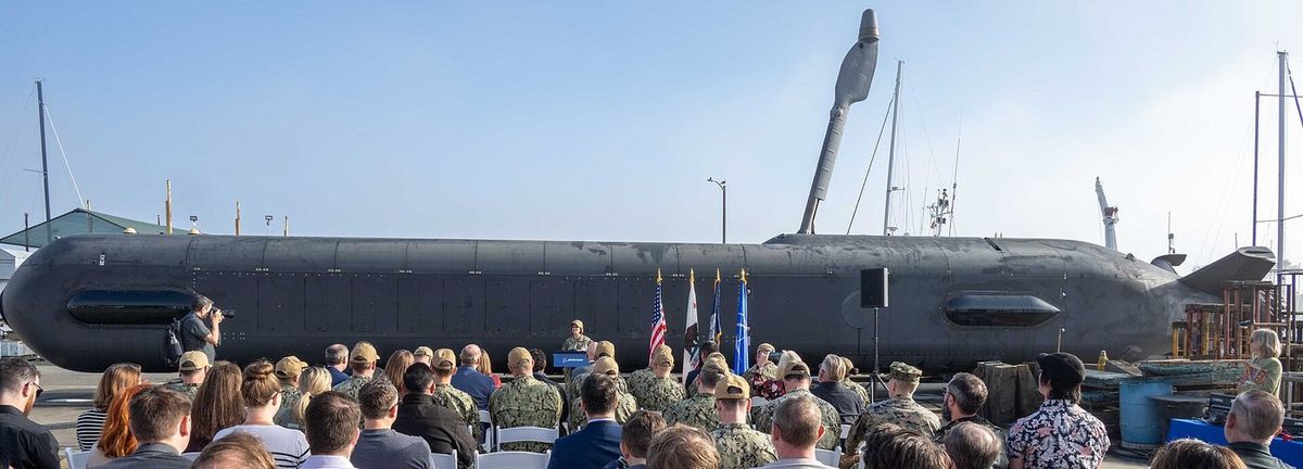 Langgestrecktes, mattschwarzes zigarrenförmiges Unterwasserfahrzeug mit hochgeklapptem Mast liegt an Land vor einem Hafen mit Segelmasten, davor ein Rednerpult mit Flaggen und Reihen von Zuhörern in Zivil und Tarnuniform