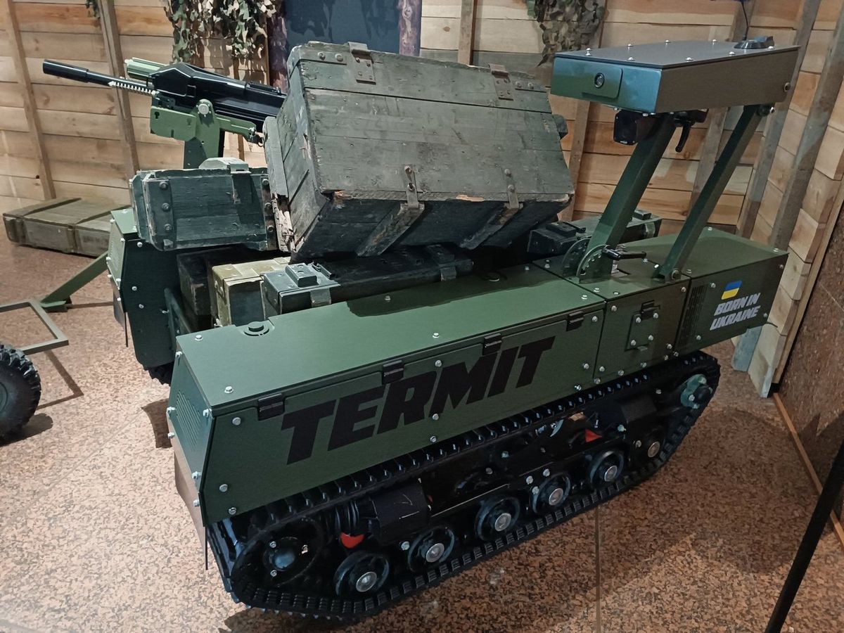 Olivgrüner Kettenroboter mit der Aufschrift TERMIT, beladen mit Munitionskisten, dahinter ein montiertes Maschinengewehr