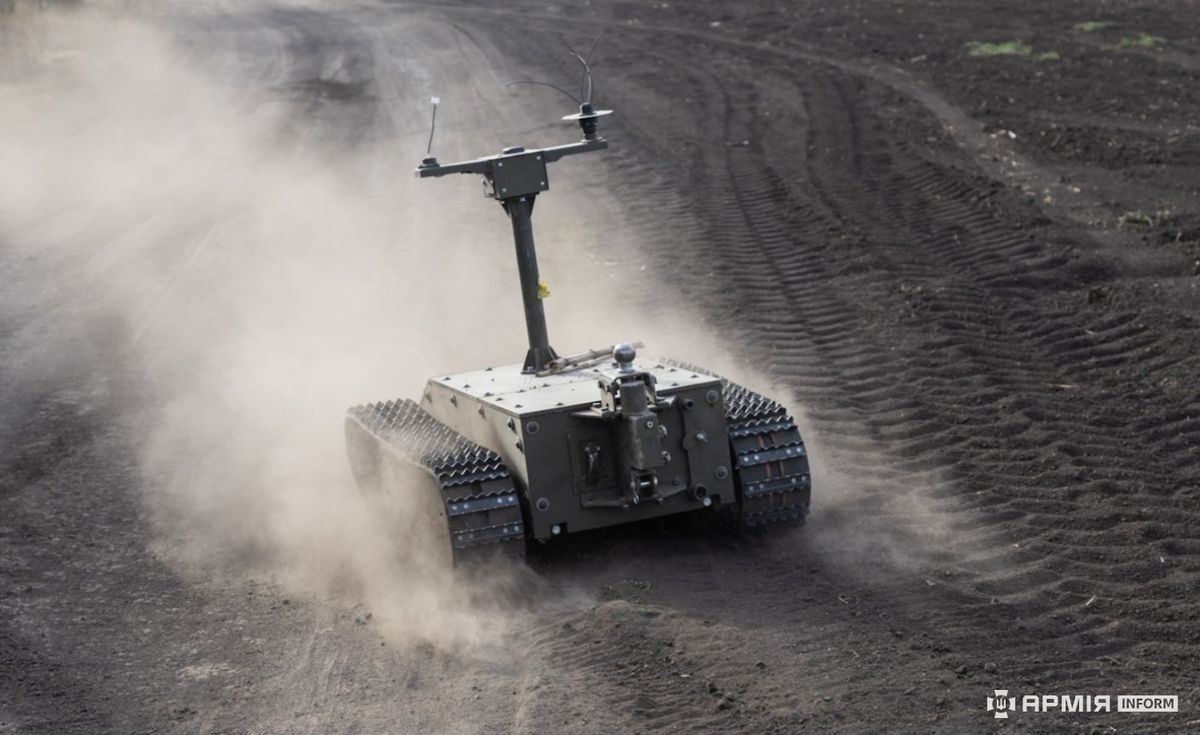 Kleiner graugrüner Kettenroboter mit hohem Mast und Antennen fährt über ein staubiges, aufgewühltes Feld und wirbelt eine Staubwolke auf, unten rechts ein Senderlogo