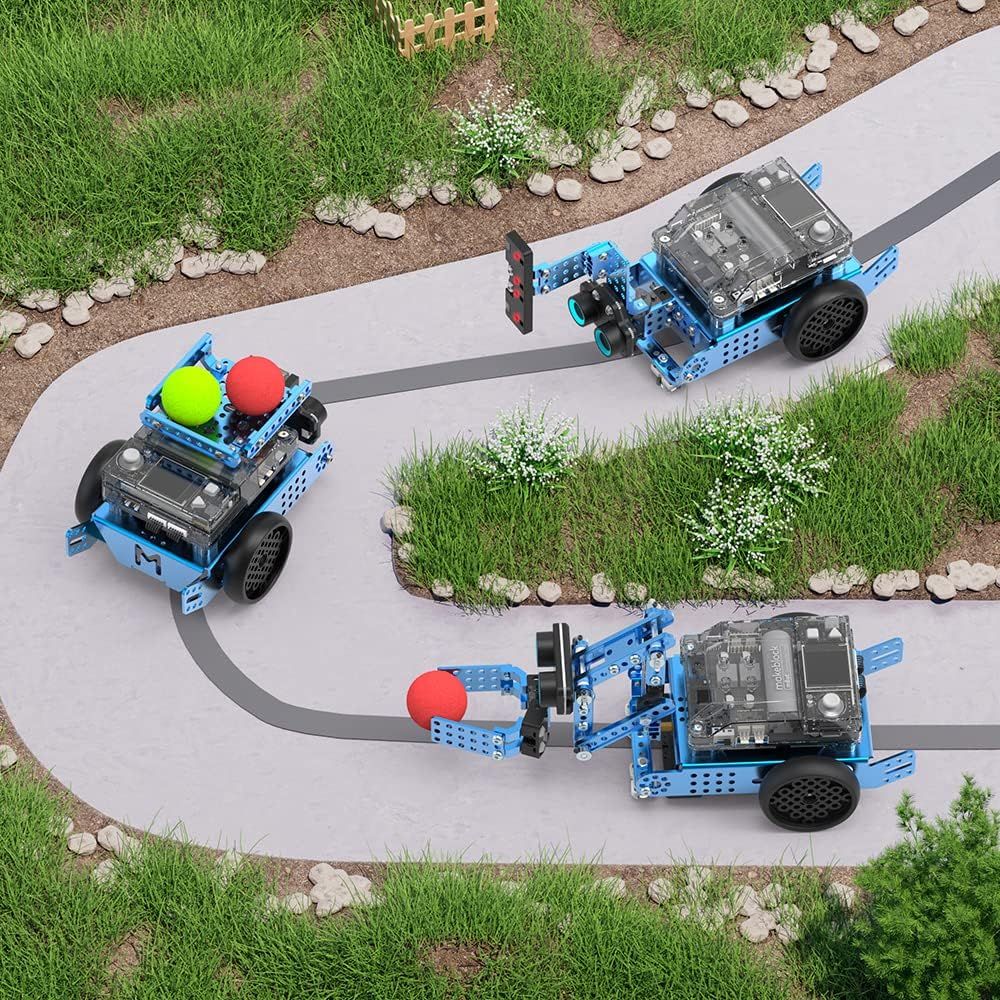 Drei blaue mBot2 auf einem grauen Weg mit schwarzer Linie zwischen Rasenflächen: oben ein Roboter mit seitlich ausgefahrenem Sensorarm, links einer mit Ladekorb voller grüner und roter Bälle, unten einer mit Greifarm, der einen roten Ball hält