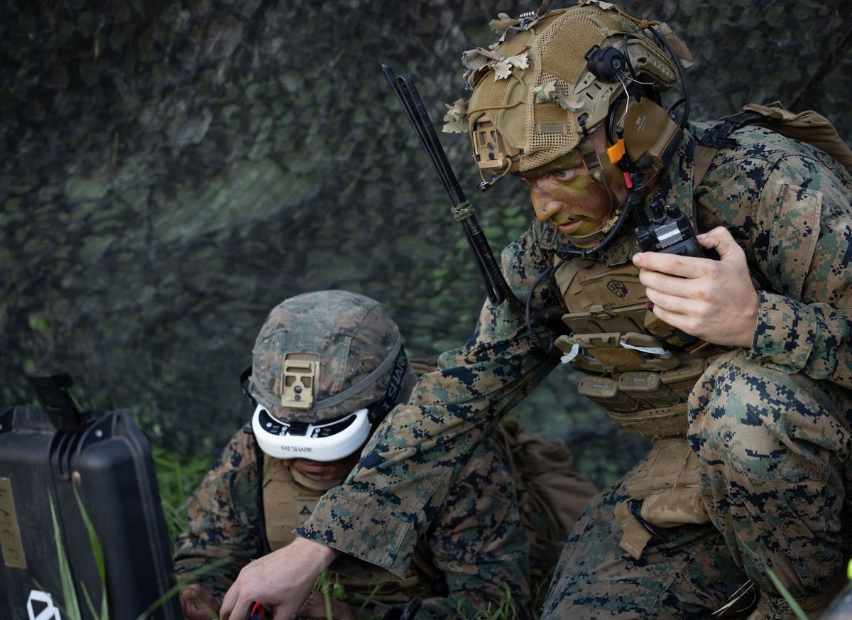 Zwei US-Marines in Tarnuniform unter einem Tarnnetz: links kniet einer mit weißer FPV-Videobrille über einem schwarzen Transportkoffer, rechts hält einer mit Tarnschminke, Helm und Headset ein Funkgerät in der Hand und blickt zur Seite
