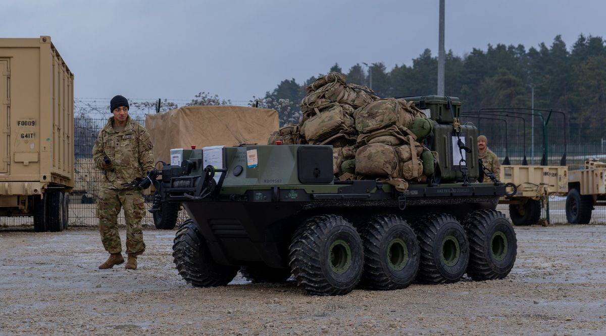 Achträdriges, dunkelgrün-schwarzes Roboterfahrzeug, hoch beladen mit Rucksäcken und Transportkisten, steht auf einem Schotterplatz zwischen Containern und Anhängern; links daneben geht ein Soldat mit Mütze und Fernsteuerung in der Hand, im Hintergrund Nadelwald