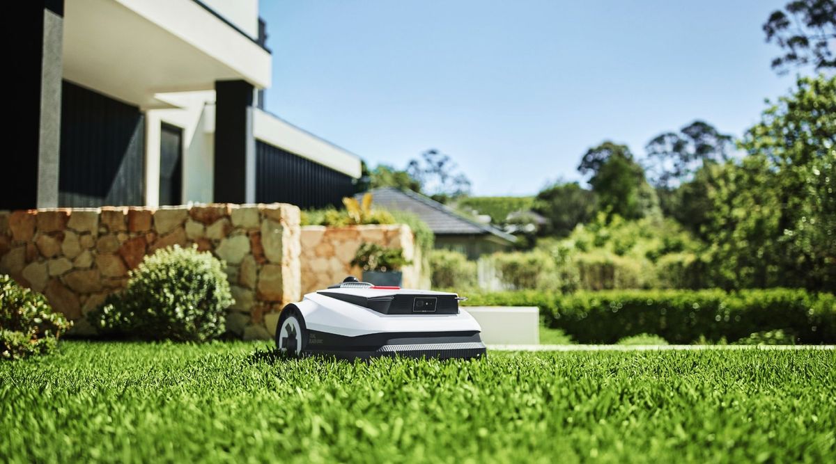 Weißer Ecovacs-Mähroboter mit schwarzer Front und Kamera fährt über einen frisch gemähten Rasen, links dahinter eine Natursteinmauer und ein modernes Haus mit dunkler Fassade, rechts Hecken und Bäume unter blauem Himmel