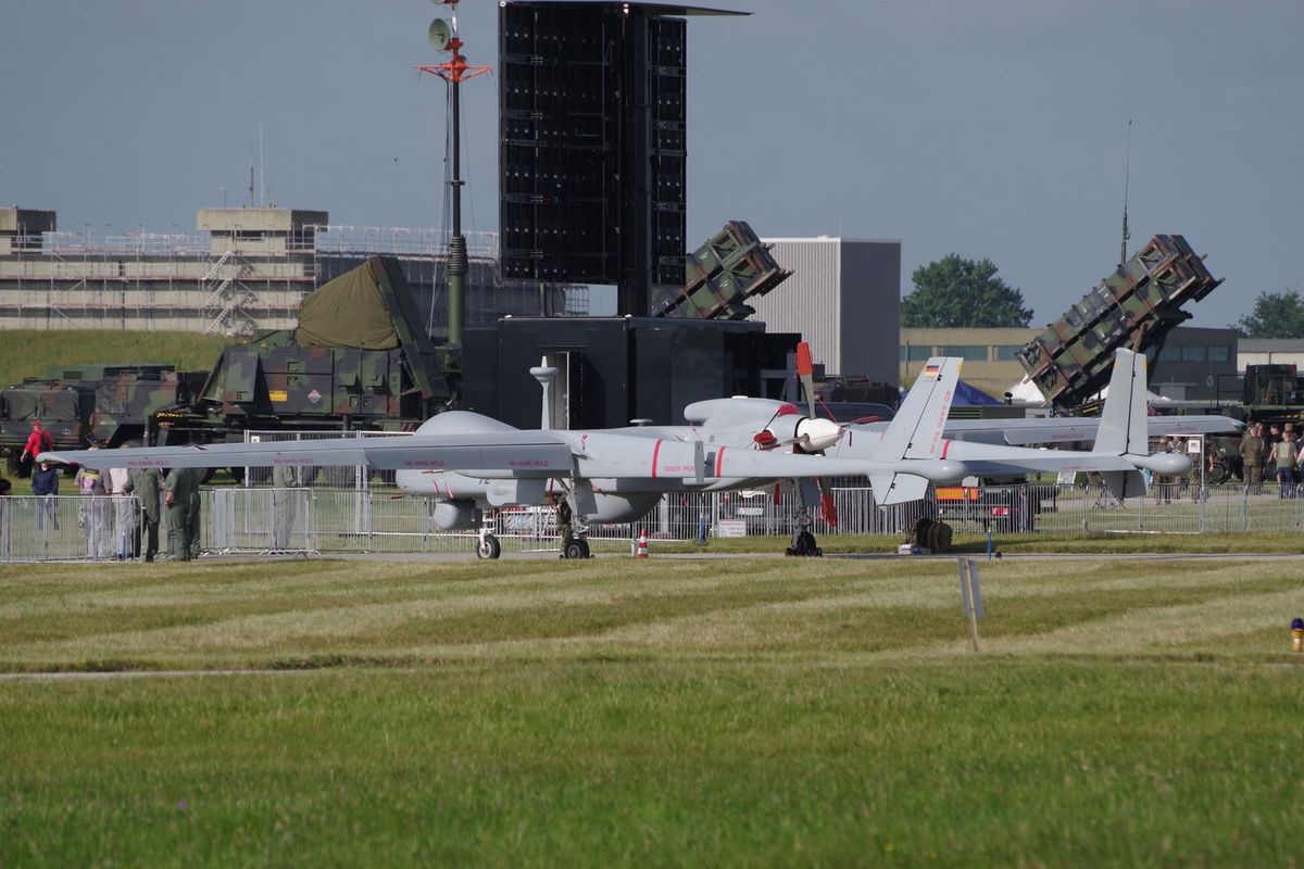 Die weiße Aufklärungsdrohne Heron TP mit deutschem Hoheitszeichen steht auf einem Flugplatz, dahinter Patriot-Startgeräte und Militärfahrzeuge
