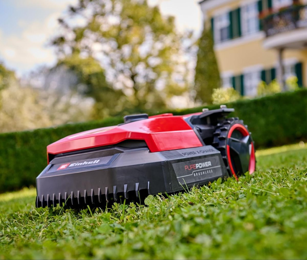 Ein roter Einhell-FREELEXO-Mähroboter der aktuellen Generation mäht bodennah fotografiert einen Rasen vor einer Hecke und einem gelben Haus.