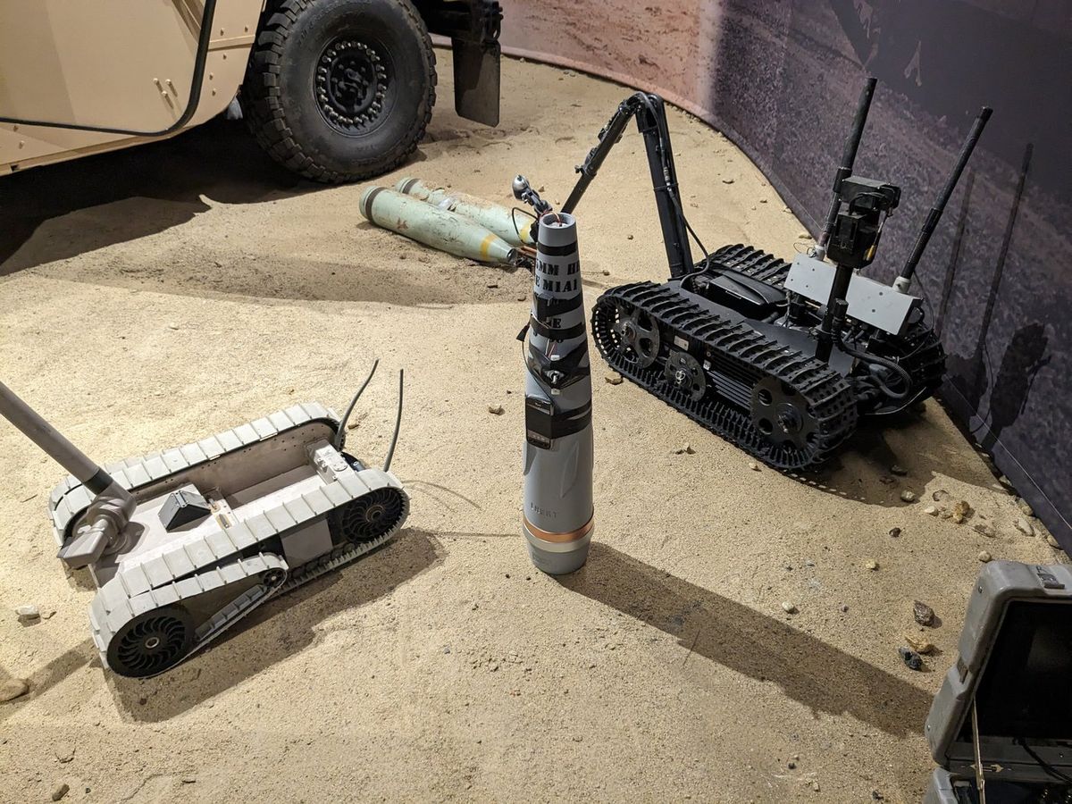 Zwei kleine Kettenroboter mit Greifarmen auf Sandboden in einer Ausstellung: links unten ein heller, flacher Roboter, rechts ein schwarzer Roboter mit aufgerichtetem Arm und Antennen, dazwischen Attrappen von Granaten, oben links das Rad eines Militärfahrzeugs