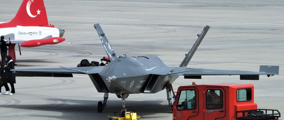 Graues pfeilförmiges Flugzeug ohne Cockpit mit nach außen geneigten Leitwerken auf einem Rollfeld, dahinter ein rot-weißes Schulflugzeug