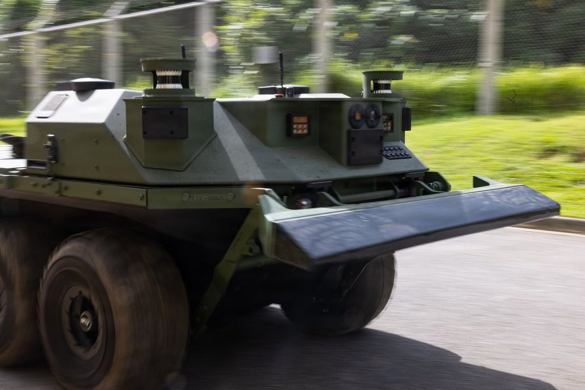 Achträdriges, olivgrünes Roboterfahrzeug mit flacher Front, Kameras und Sensoraufbauten fährt auf einer Straße an einem Zaun und Grünflächen vorbei, der Hintergrund ist durch die Bewegung verwischt