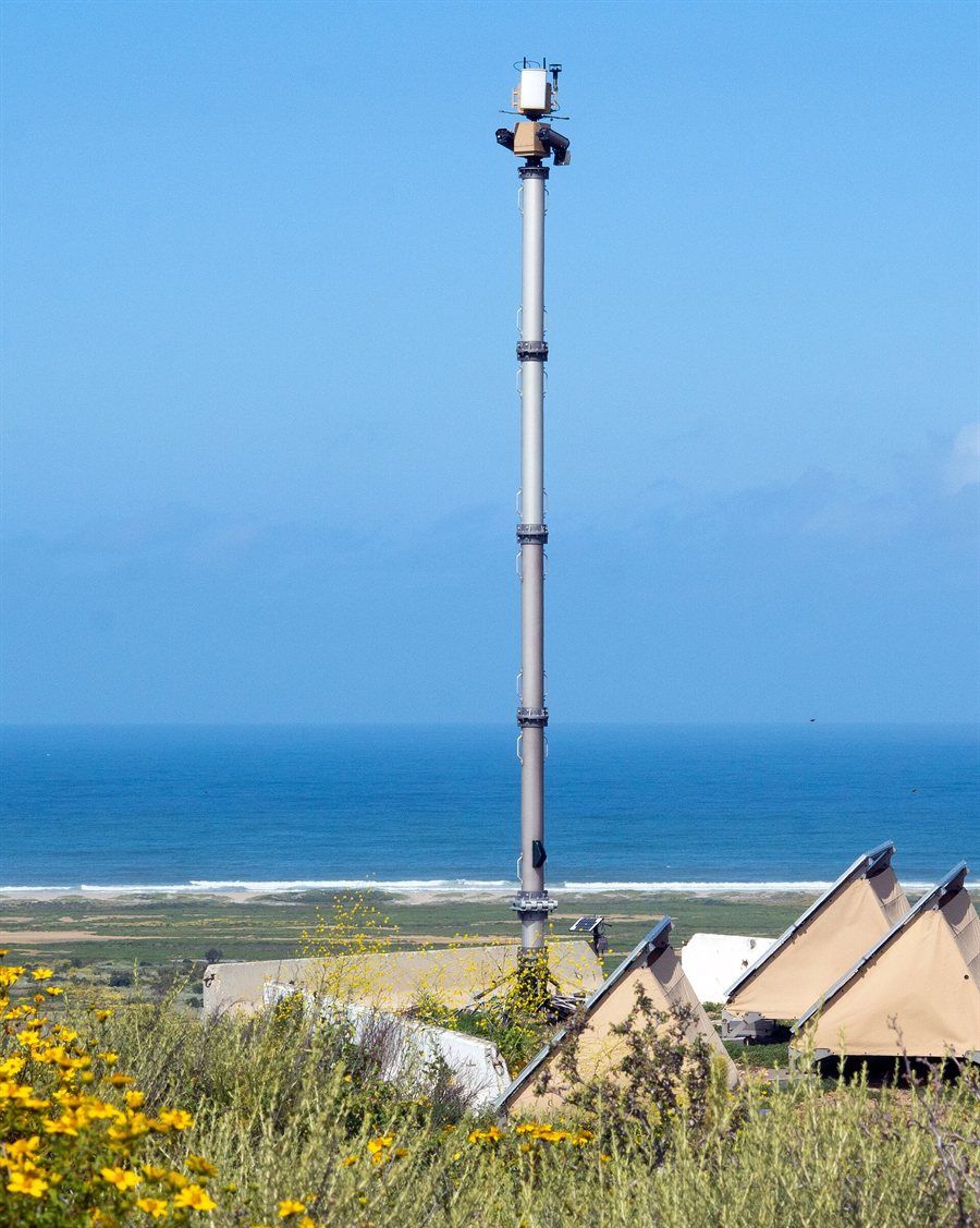 Hoher schlanker Mast mit Kamera- und Radarkopf an der Spitze steht auf einer Blumenwiese über der Pazifikküste, daneben beigefarbene Zelte