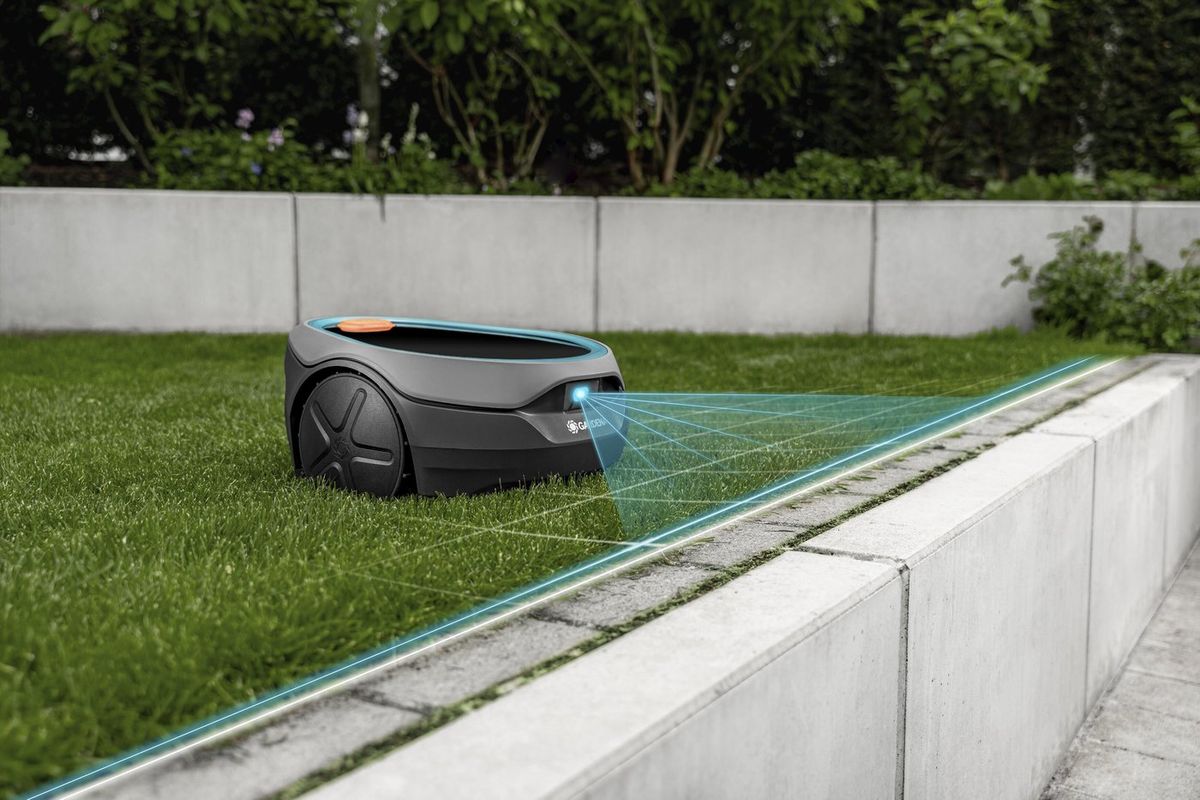 Flacher grauer Mähroboter auf einem Rasen vor einer Betonmauer, eine eingeblendete türkisfarbene Grafik zeigt, wie seine Frontkamera die Kante zu einem Plattenweg erfasst