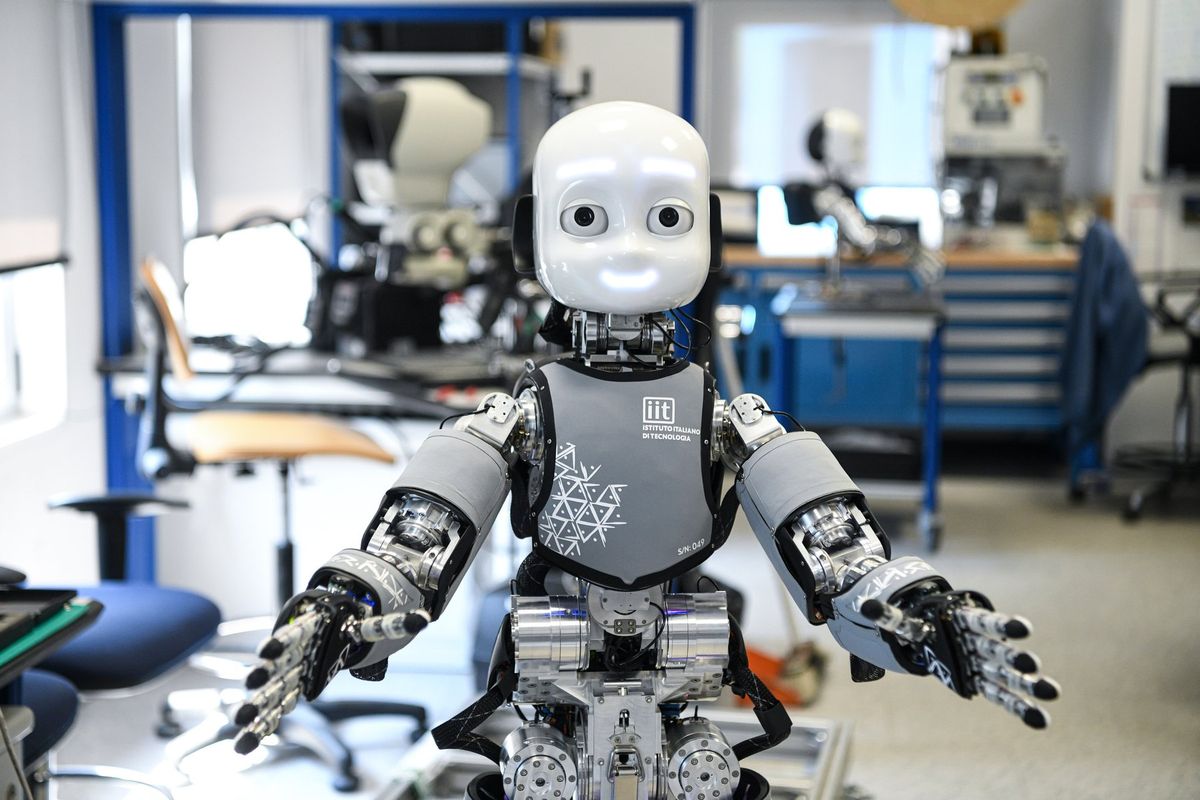 Humanoider Forschungsroboter iCub mit weißem Kopf, leuchtend weißen Augenbrauen und lächelndem Mund, grauer Textilhülle mit iit-Logo und der Aufschrift S/N: 049 auf der Brust, beide Arme mit metallischen Fingern seitlich ausgebreitet; dahinter ein Labor mit blauen Regalrahmen, einem orangefarbenen Drehstuhl links, blauen Schubladenschränken rechts und einem zweiten, unverkleideten Roboter im Hintergrund