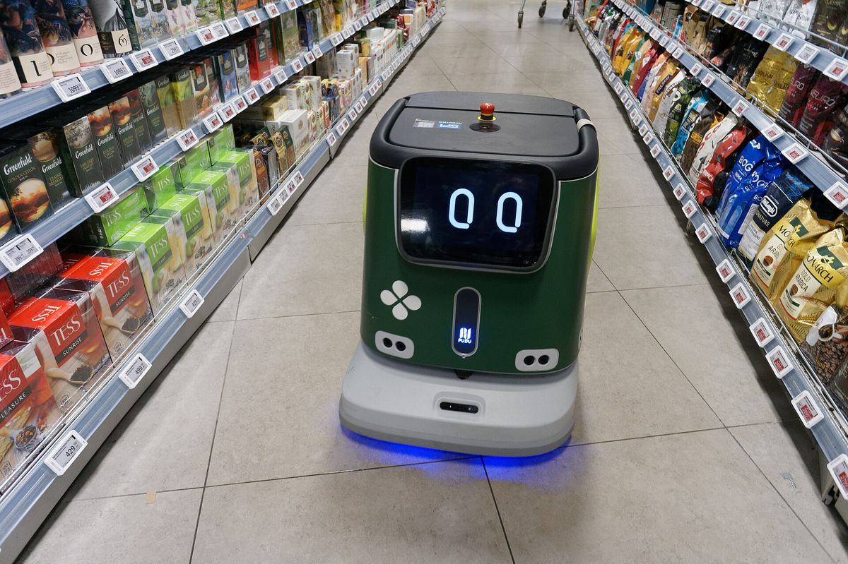 Reinigungsroboter Pudu CC1 mit Display-Augen fährt frontal durch einen Supermarktgang zwischen Tee- und Kaffeeregalen, unter dem Gehäuse leuchtet ein blaues Lichtband.
