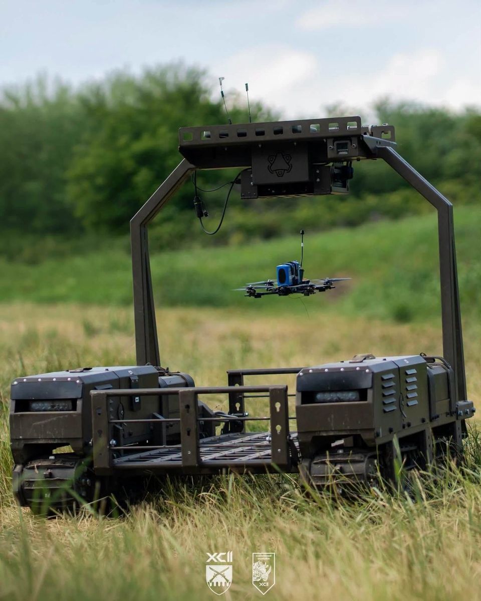 Kettenroboter auf einer Wiese mit einem Startgestell über der Ladefläche, darunter schwebt eine blaue FPV-Drohne