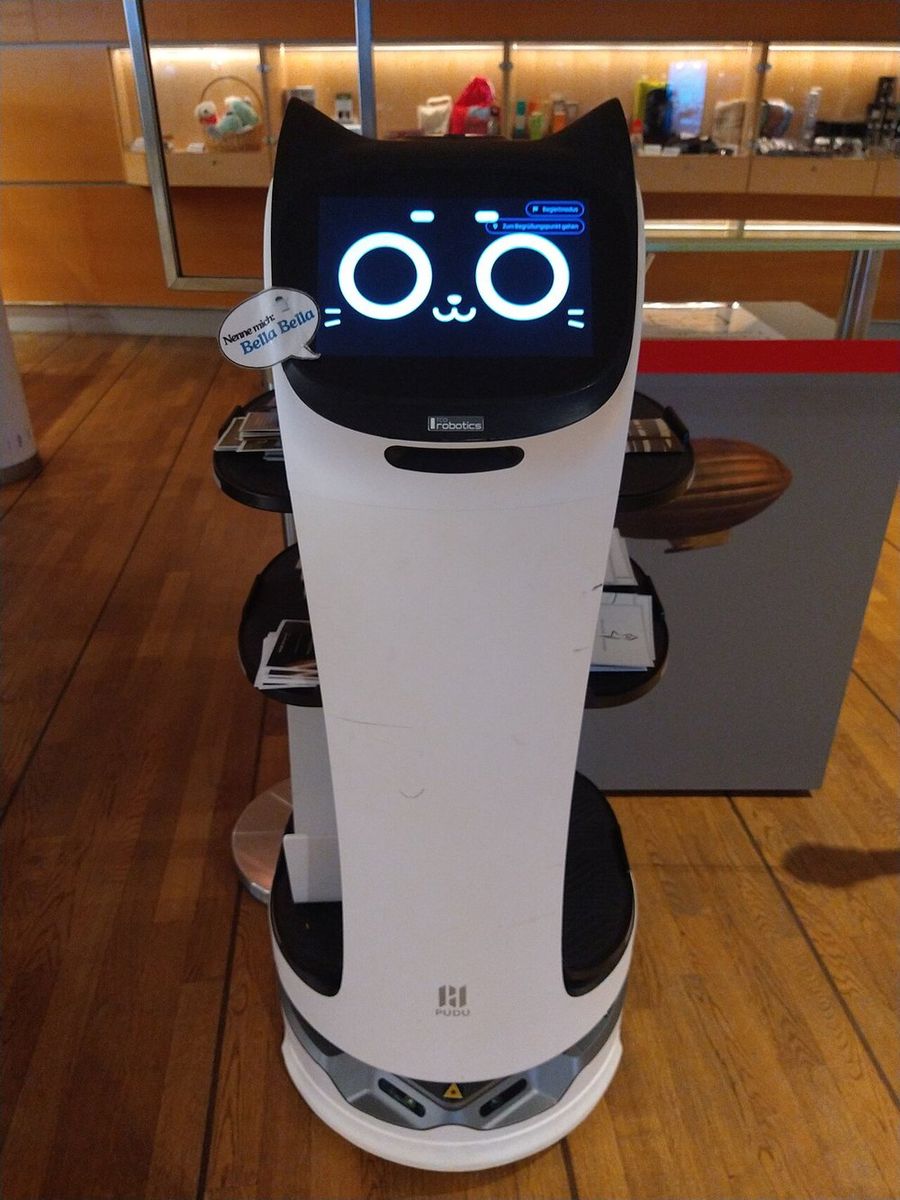 Weißer Roboter mit schwarzem Katzenkopf und Augen auf dem Display, seitlich drei schwarze Tablare mit Prospekten, am Kopf ein Aufkleber „Nenne mich: Bella Bella“, unten das Pudu-Logo, er steht auf Holzparkett vor einer beleuchteten Vitrine